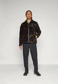 Giacca in pile nera con motivi astratti gialli e bianchi, con zip frontale completa, colletto alto, indossata con pantaloni scuri e stivali neri.