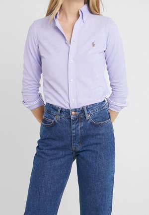 Vrouw die een lichtpaarse blouse met knopen draagt met opgerolde mouwen en een high-waisted blauwe spijkerbroek, staand voor een effen achtergrond.