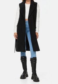 Long gilet noir avec un col cranté, des fentes à l'avant et une taille à nouer. Porté par-dessus un pull court blanc et un jean bleu clair, associé à des bottes noires à lacets.