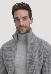Grauer, gestrickter Reißverschluss-Pullover mit einem hohen Kragen, der ein strukturiertes Muster aufweist und darunter ein glattes, graues Langarmshirt trägt.