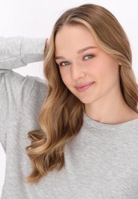 Ljusgrå långärmad sweatshirt med rund hals, med en mjuk textur. En person med vågigt, ljusbrunt hår poserar med en hand på huvudet.