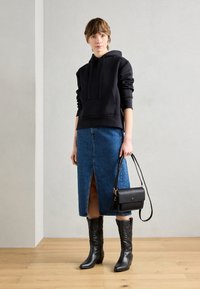 Hoodie noir avec poche kangourou, jupe en denim bleu avec fente avant, bottes noires en cuir jusqu'au genou et un petit sac bandoulière noir.