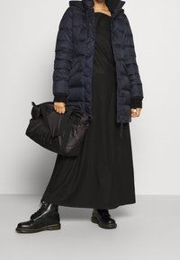 Gewatteerde marineblauwe jas met capuchon, lange zwarte jurk, zwarte veterschoenen en een zwarte tas met stoffen hengsels. Even textuur, minimalistisch design.