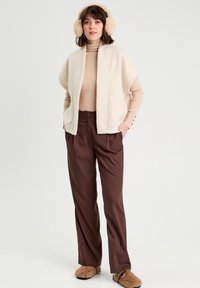 Un gilet beige duveteux sur un col roulé marron à côtes, associé à un pantalon ample marron et des chaussons en fausse fourrure beige. Des cache-oreilles complètent le look.