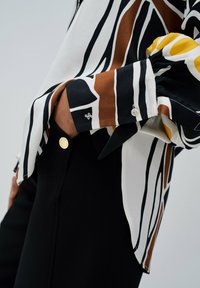 Chemise à manches longues présentant des motifs abstraits audacieux en noir, blanc, marron et jaune, avec des boutons aux poignets et une texture lisse.