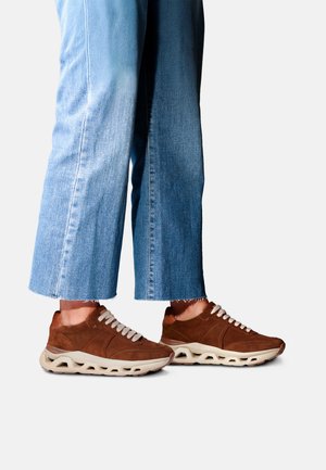 Personne portant un jean bleu court et des baskets marron avec des semelles et des lacets blancs, debout sur un fond blanc.