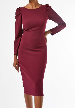 Femme portant une robe midi bordeaux ajustée et texturée à manches longues avec des épaules bouffantes, posant avec une main sur la hanche.