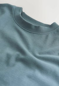Tricou din bumbac de culoare albastră deschisă cu guler rotund. Prezintă o textură netedă și detalii cusute subtile de-a lungul gulerului.