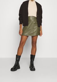 Cardigan marron à côtes, haut beige ajusté, jupe en simili cuir olive avec une fente à l'avant, et bottines noires à semelle épaisse.
