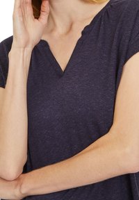 Tmavě námořnická modrá halenka s výstřihem do V a krátkým rukávem, měkkou texturou a volným střihem. V látce je viditelný jemný melírovaný vzor.