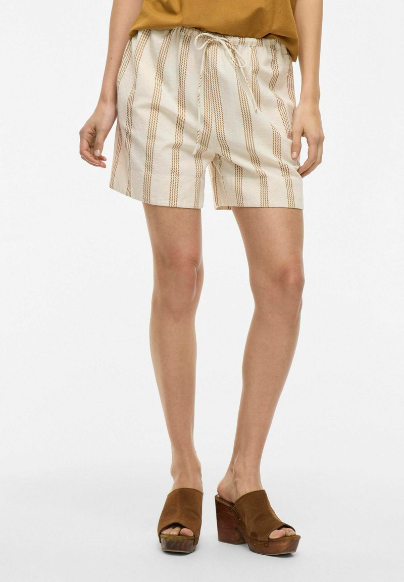 Pantalones cortos de color beige claro con rayas verticales en marrón y naranja. Cintura elástica con cordón. Llevados con zuecos marrones.