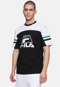 Svart och vit t-shirt med kort ärm och gröna randiga detaljer, samt en framträdande FILA-logotyp på framsidan. Tillverkad av bomullstyg.
