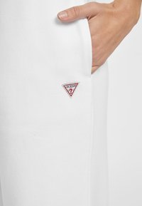 Bílé tepláky vyrobené z měkkého materiálu s malým červeným a bílým trojúhelníkovým logem s nápisem "GUESS" blízko kapsy.