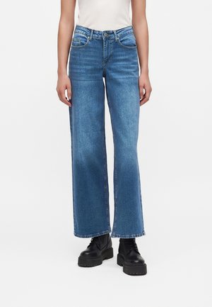 Personne portant un jean large bleu moyen et des bottes noires épaisses à lacets, debout devant un fond blanc uni.