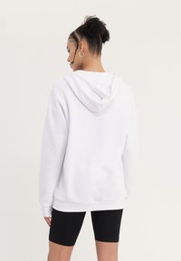 Felpa bianca con cappuccio e vestibilità ampia, maniche con polsini a costine, abbinata a pantaloni biker neri. Tessuto liscio, senza motivi o dettagli visibili.