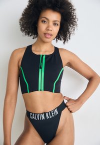 Čierny športový podprsenka s zelenými zipsovými detailmi a bočnými akcentmi, v kombinácii s čiernymi bikiny nohavicami s vysokým pásom, na ktorých je branding "CALVIN KLEIN".