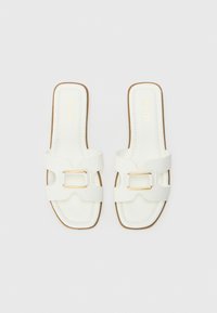 White slide sandals with two wide straps featuring a gold buckle, flat sole, and stitched detailing along the edges. Brand name embossed in gold.
