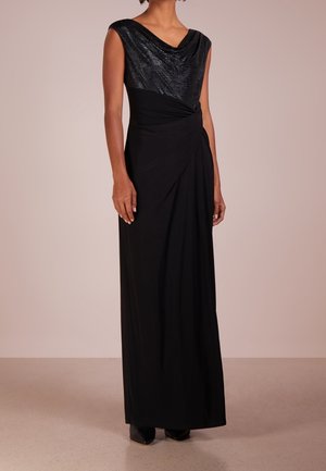 Nainen, joka pitää mustaa hihiatonta iltapukua, jossa on teksturoitu yläosa ja laskostettu vyötärö, seisomassa neutraalilla taustalla.