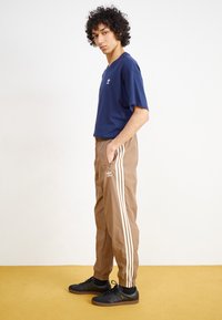 Camiseta azul-marinho de manga curta, combinada com calças castanhas de cintura elástica com riscas brancas laterais e um logótipo bordado. Ténis pretos.