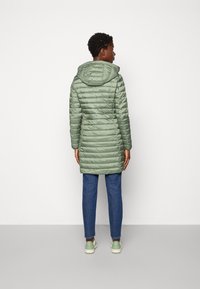 Gefütterter, langer olivgrüner Mantel mit Kapuze, der eine gesteppte Textur und elastische Bündchen aufweist. Kombiniert mit blauen Jeans und hellgrünen Sneakern.