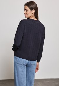 Navy gestrickter Pullover mit einem strukturierten Fischgrätmuster, lockerer Passform, überschnittenen Schultern und gerippten Bündchen, kombiniert mit hellblauenweiten Jeans.