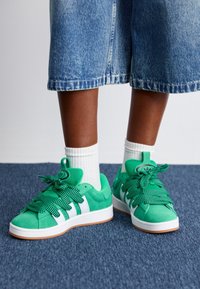 Gröna mockaskor med tjocka mönstrade gröna snören, vita ränder och en rund logotyp. Kombination med vita ankelsockor och blåjeansshorts.