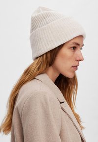 Berretto beige a coste con una parte superiore a punta, realizzato in morbido tessuto a maglia. Indossato con un cappotto beige, che mostra una sottile texture.