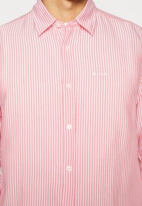 Homme portant une chemise à rayures verticales roses et blanches, boutonnée avec col, et le logo "BOSS" brodé sur la poitrine.