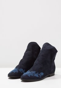 Botines de ante en azul marino con punta afilada y suela plana, adornados con detalles de rhinestones azules en la parte delantera.