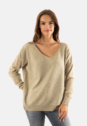 Maglione beige lavorato a maglia con scollatura a V, vestibilità ampia, maniche lunghe e orlo a coste. Indossato con jeans grigi, mostrando una consistenza morbida.