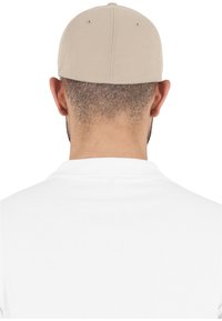 Beige Baseballkappe mit strukturierter Krone und belüftenden Ösen, rückwärts getragen. Der Kragen eines weißen Poloshirts ist sichtbar.