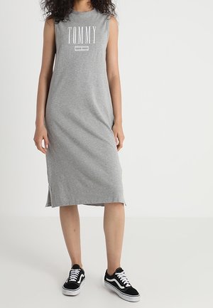 Robe grise sans manches avec inscription "TOMMY" en blanc. Matière douce en coton, longueur au genou avec des fentes latérales. Portée avec des baskets noires.