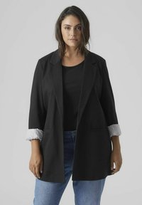 Blazer noir à manches longues, retrouss�ées pour révéler une doublure à rayures blanches. Porté sur un haut noir, associé à un jean bleu. Deux poches avant.