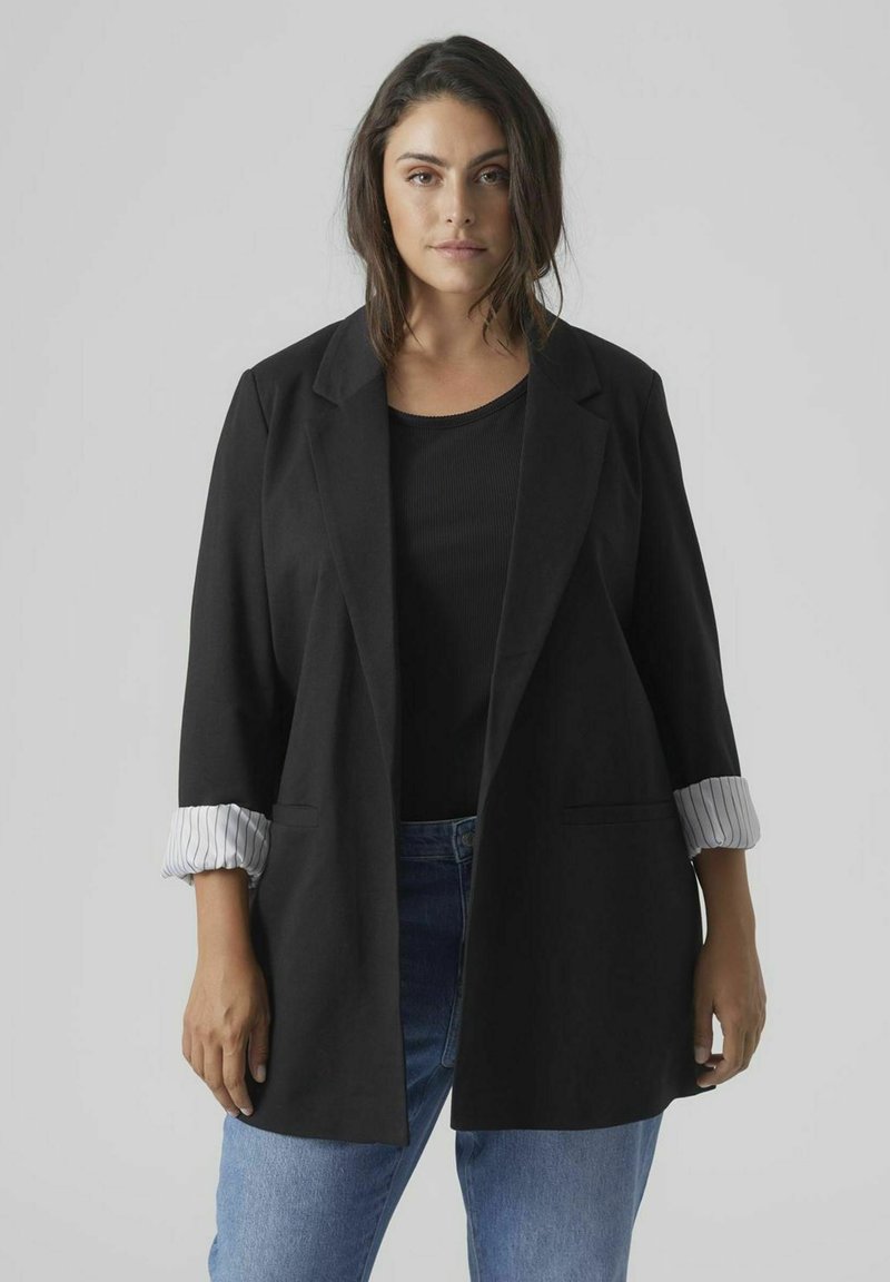 Blazer noir à manches longues, retroussées pour révéler une doublure à rayures blanches. Porté sur un haut noir, associé à un jean bleu. Deux poches avant.