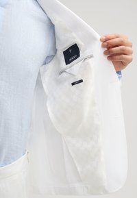 Weißer Blazer mit glatter Textur und gemustertem Futter. Das Innenlabel trägt die Aufschriften "JOOP!" und "SLIM FIT." Eine Hand hält den Blazer geöffnet.