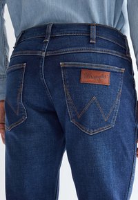 Detail osoby nosiacej tmavomodré rifle Wrangler, zobrazujúci zadné vrecká a koženú značku na pravom vrecku.