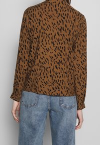 Braune Bluse mit einem schwarzen Tierdruck-Muster, lockere Passform, lange Ärmel und Kragen, kombiniert mit hellblauen Jeans.