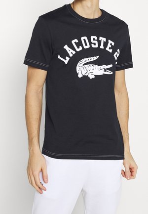Homme portant un t-shirt noir à manches courtes avec le texte blanc "Lacoste" et le logo crocodile, associé à un pantalon blanc sur un fond uni.