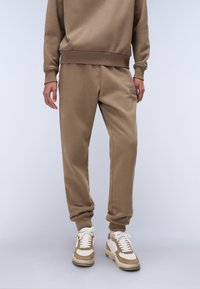 Bruine katoenen sweatshirt en bijpassende joggers, met ribgebreide boorden en een subtiele logo-detail op de zak. Sneakers met beige accenten.