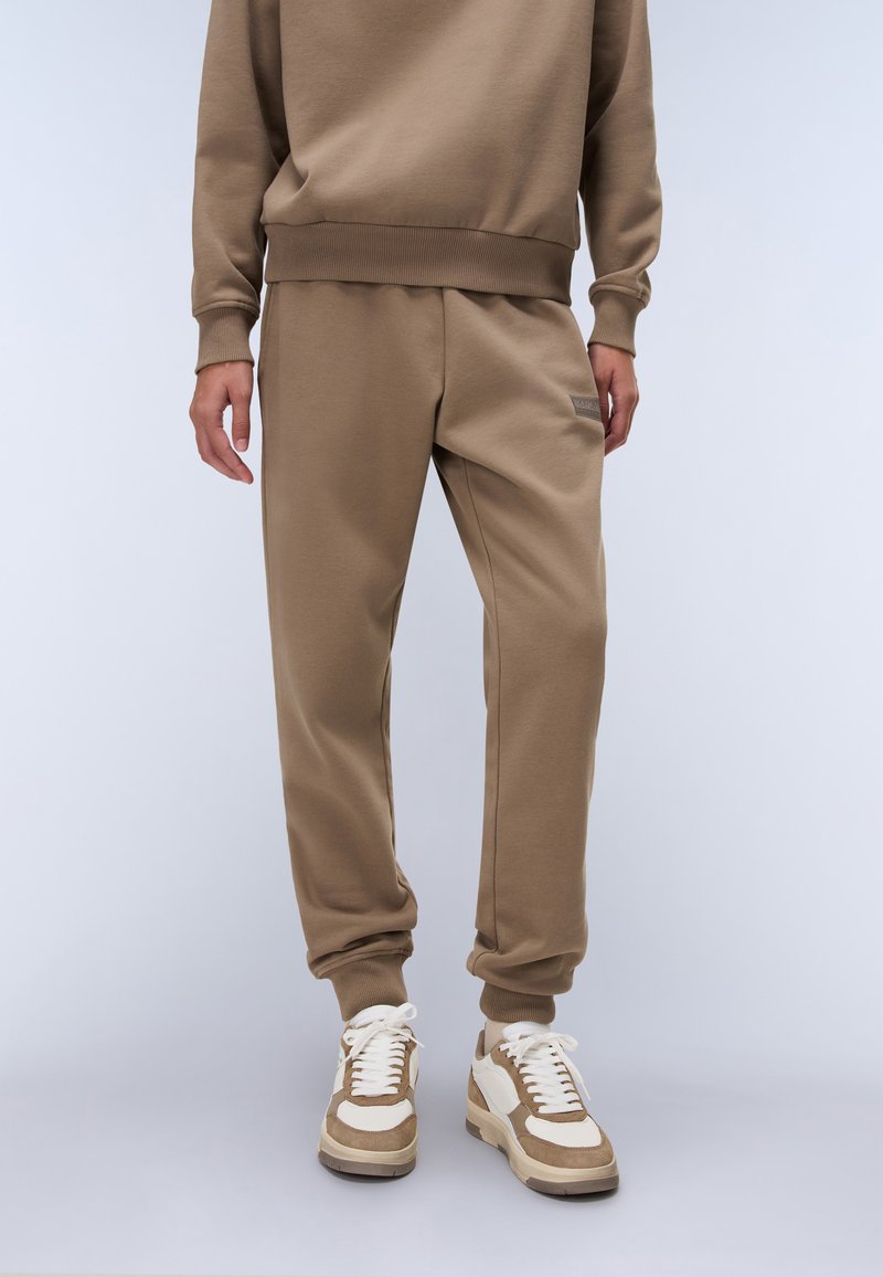 Bruine katoenen sweatshirt en bijpassende joggers, met ribgebreide boorden en een subtiele logo-detail op de zak. Sneakers met beige accenten.