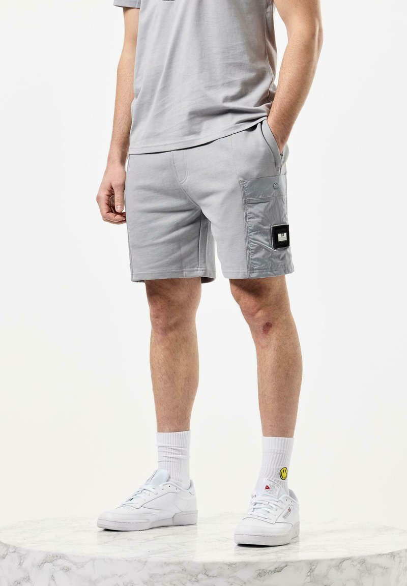 Grå cargo shorts med en sidelomme, laget av et mykt materiale. Brukt med hvite sneakers og ribbestrikkede sokker, med et smiley-logo.
