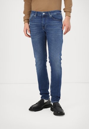 Person wearing slim-fit blue jeans, black loafers, white socks, and a brown waffle-knit sweater, standing against a plain white background.