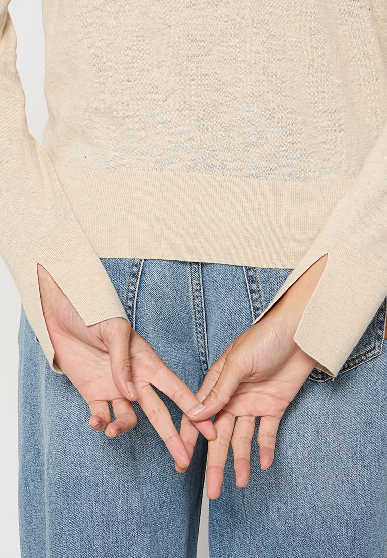 Beżowy sweter dzianinowy z długimi rękawami, z bocznymi rozcięciami na mankietach. Noszony z jasnoniebieskimi dżinsami typu high-waist. Ręce skrzyżowane za plecami.