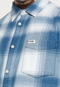 Chemise à carreaux bleue et blanche avec un col boutonné, dotée d'une poche et d'une petite étiquette de marque. Fabriquée en tissu léger.