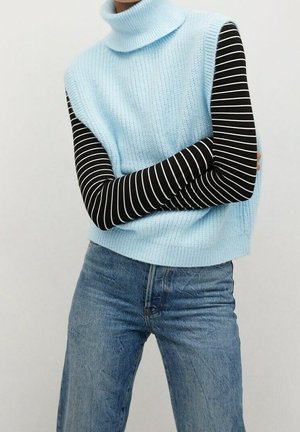 Osoba nosící světle modrý bez rukávů rolák přes černobíle pruhované tričko s dlouhým rukávem a modré džíny, s rukama zkříženýma na prsou.