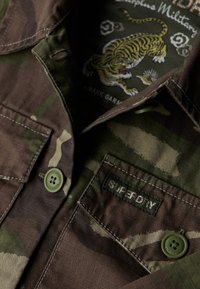Koszula w stylu militarnym w kamuflażowym wzorze w kolorze brązowym i zielonym, z kołnierzykiem, dwoma kieszeniami na piersi oraz zielonym guzikiem z napisem "Superdry".