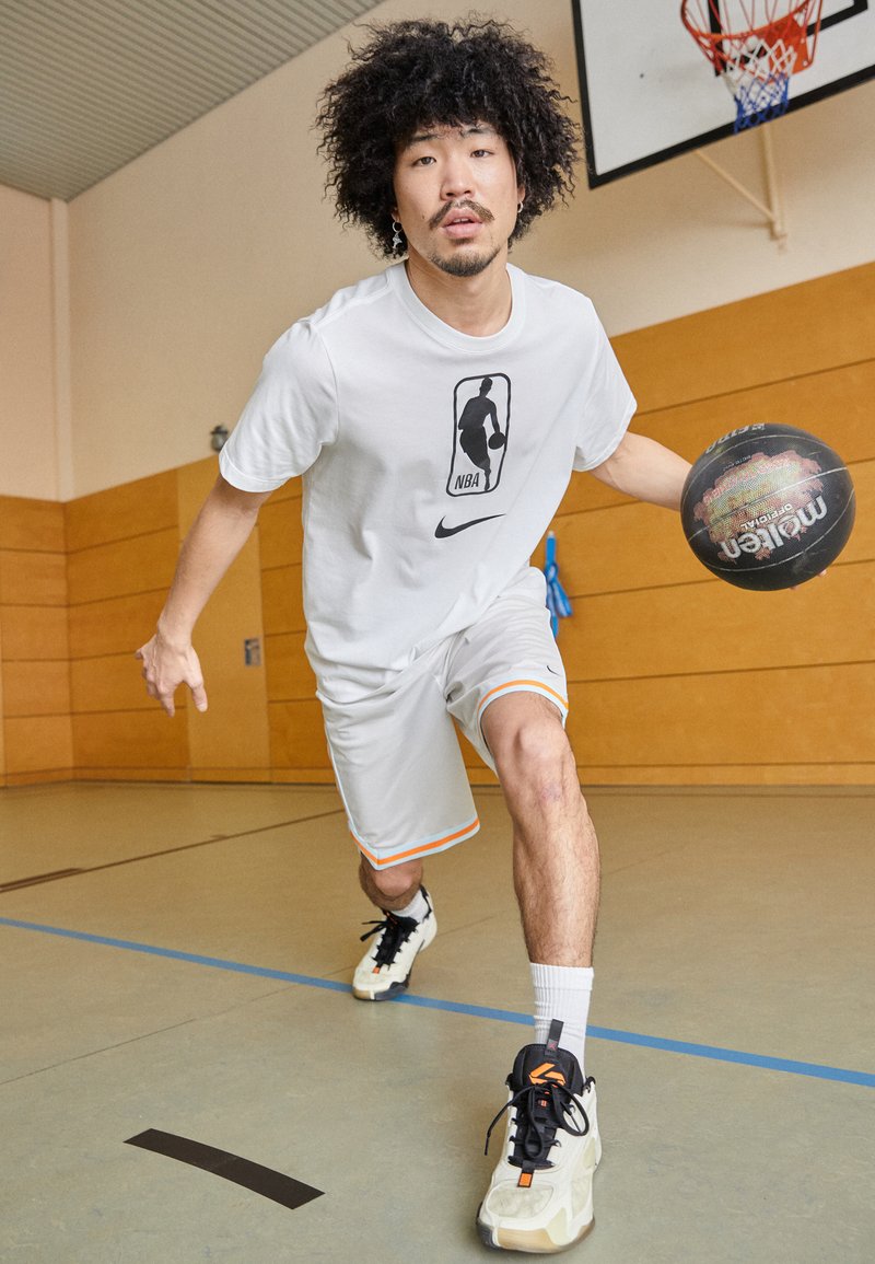 Jugador de baloncesto con una camiseta blanca del logo de la NBA y pantalones cortos grises con ribete naranja, driblando un balón de baloncesto negro en un gimnasio.