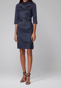 Granatowa sukienka koszulowa z wykałaczanego kołnierzem, rękawami o długości trzech czwartych, talią z paskiem i prostym dołem. W zestawie z czarnymi sandałami na obcasie.