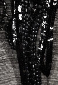 Czarne cekinowe zdobienia na teksturowanej szarej tkaninie, z dyskowatymi i płaskimi kształtami w różnych rozmiarach, ułożonymi w warstwowe pasma.