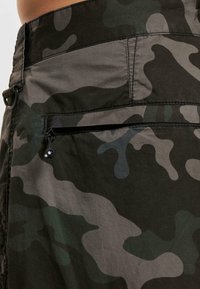Närbild av byxficka i camouflage-mönster med en svart dragkedja och ett litet tygögl med en metallring på midjebandet.